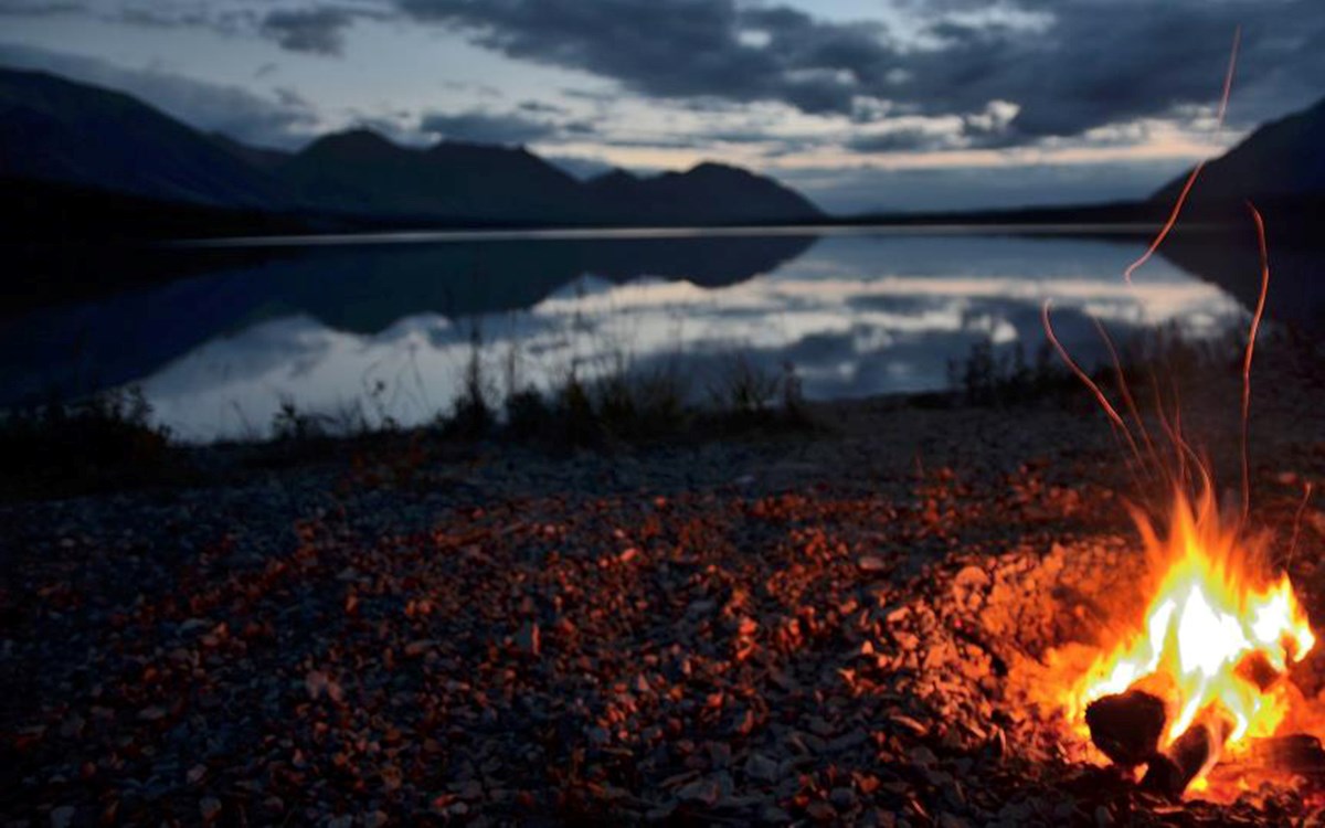 A campfire burns on a beach in the right, bottom corner of the photo with a lake and mountains at dusk in the background.