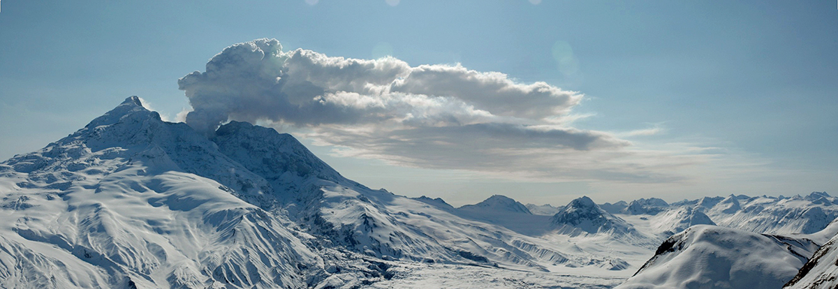 A plume of steam rises from a snow and ice-capped volcano on a clear day.