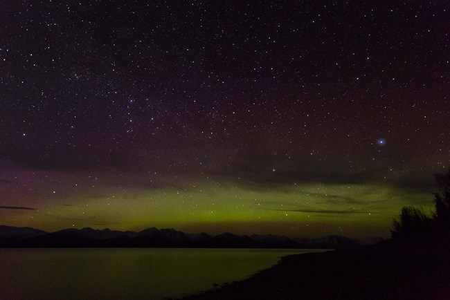A night sky is lit up with hundred of stars. On the horizon, a green light, the aurora, cuts across the sky