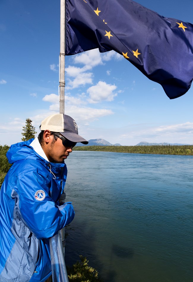 a man stands on a wooden tower counting fish from above next to an Alaska state flag