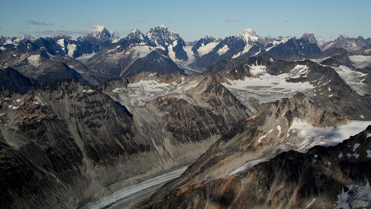 aerial view of jagged mountains with snow patches and multiple alpine glaciers flowing down their flanks and in-between them.