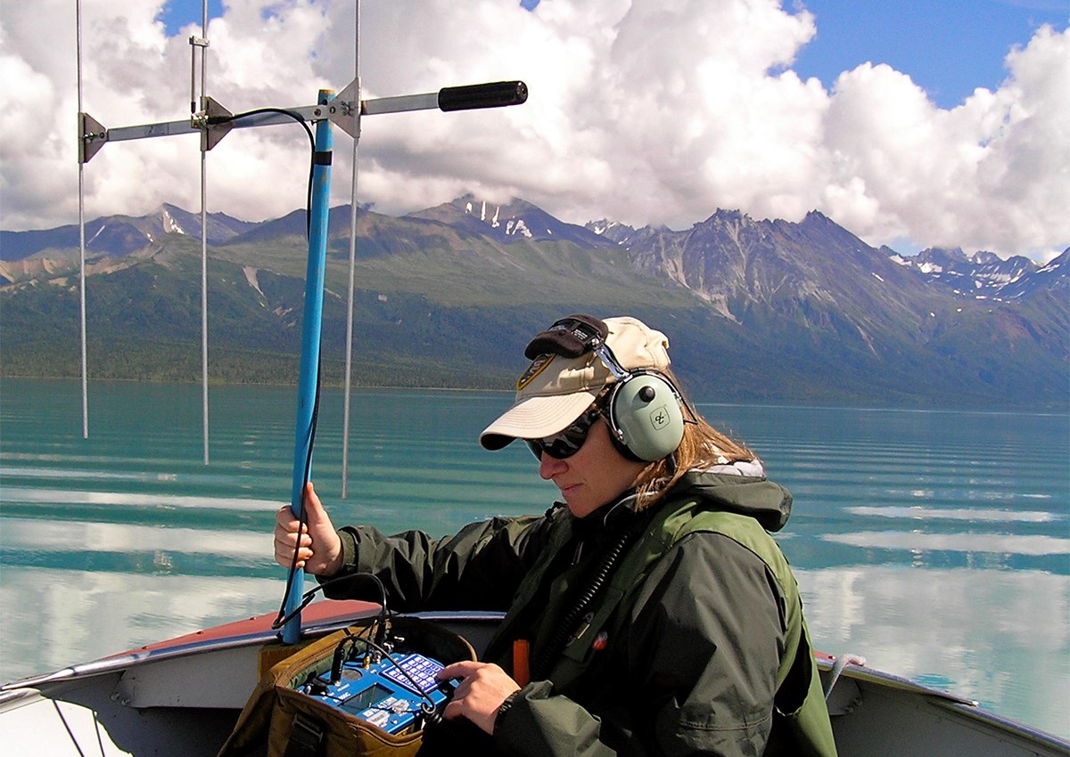 Woman holding a large antenna sits in a boat on a lake surrounded by colorful mountains.