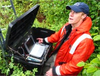man in an orange rain coat sets up a computer and other equipment outdoors