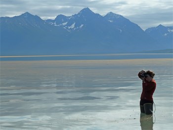 person in hip waders standing in a lake, taking a picture; mountains in the distance
