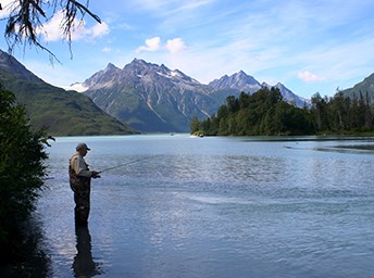 man fishing in a lake, surrounded by forest and tall mountains