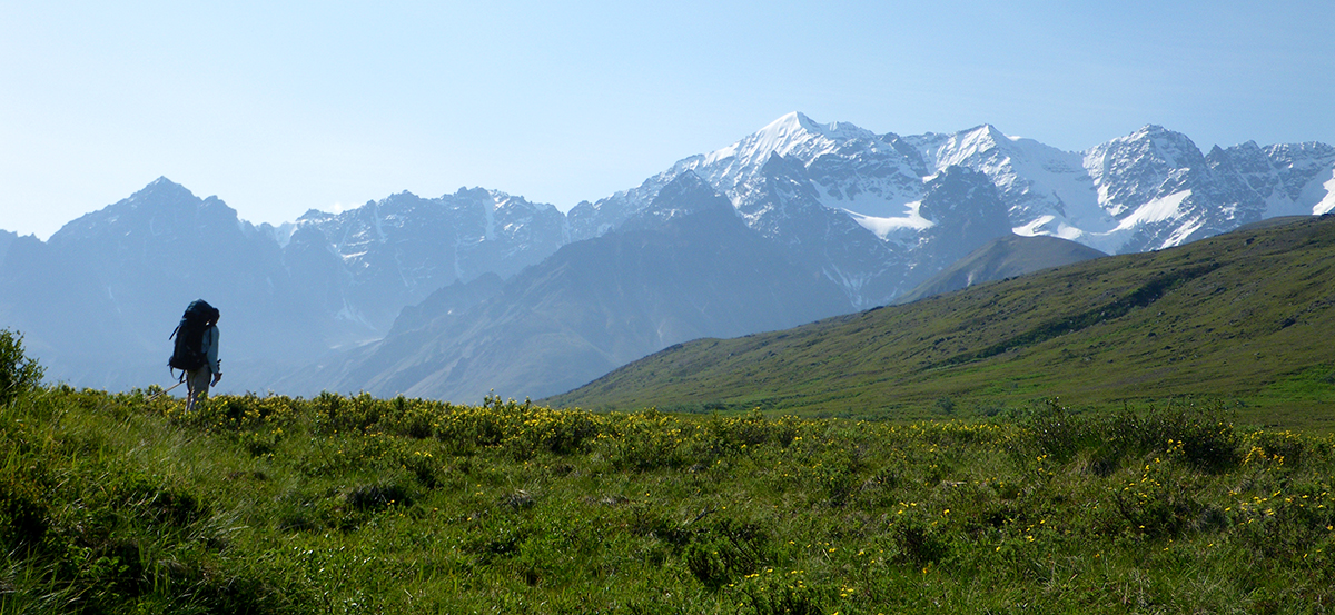 Photo of a backpacker hiking across tree-less hills with tall, snow-capped mountains in the distance.