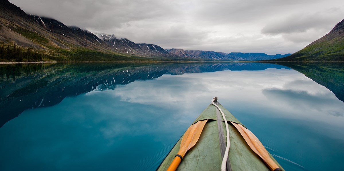 looking over the bow of a kayak across a blue lake toward cloud-covered mountains
