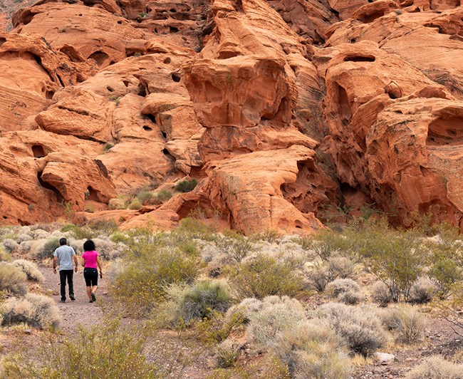 Two hikers walking next to giant red rocks.
