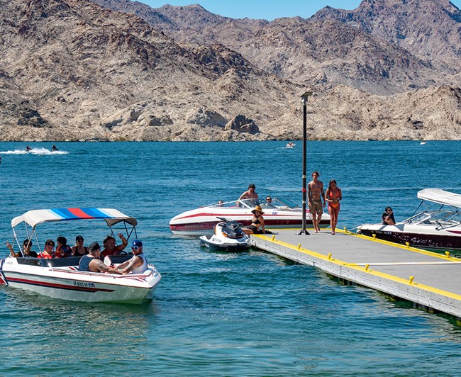 Several boats and water crafts next to a pier with multiple people in swimwear on it.