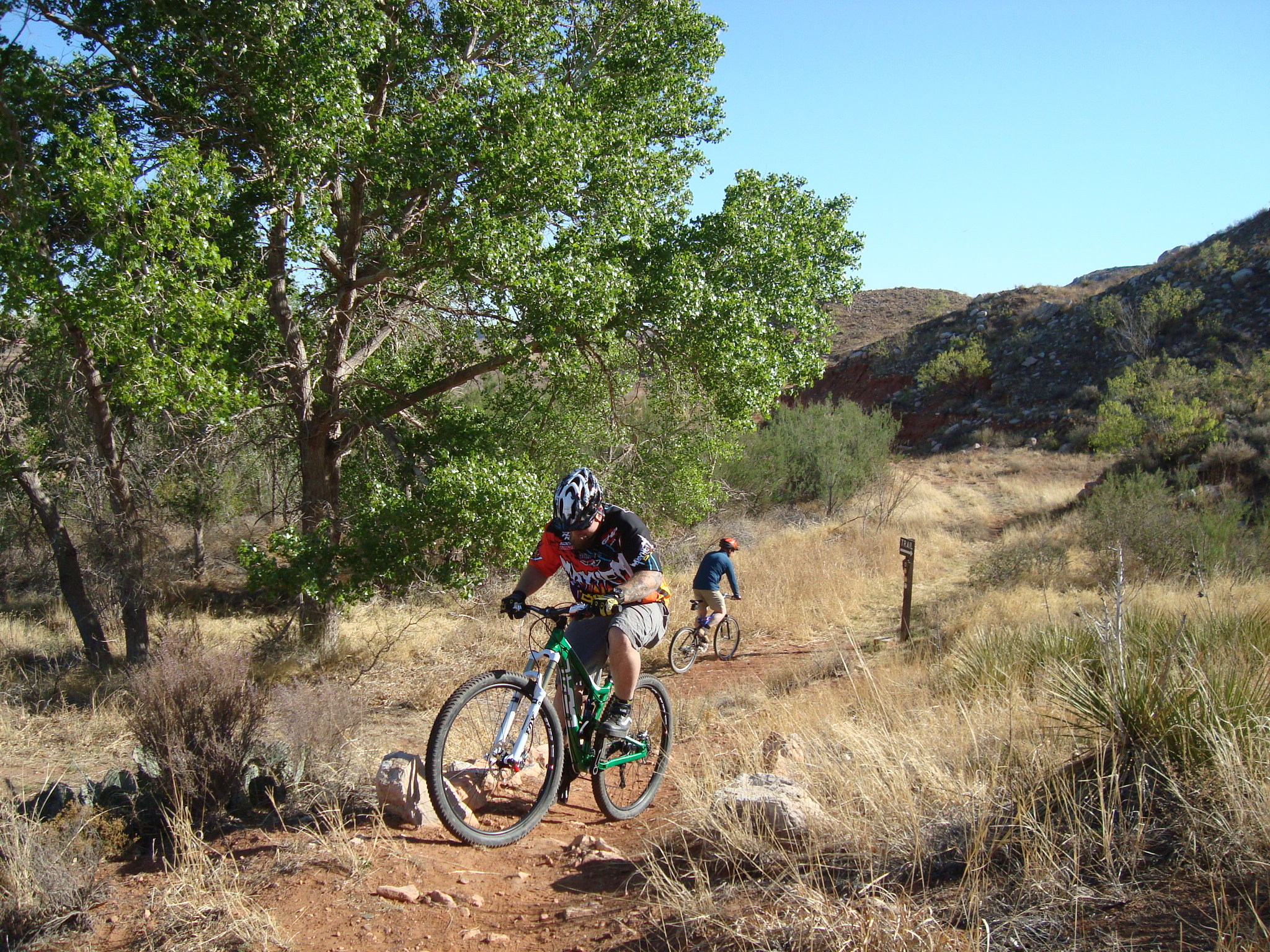 Two mountain bikers at Lake Meredith biking along the Harbor Bay Trail.  There are green cottonwoods and grasses along the trail.  It is a sunny day with blue skies.