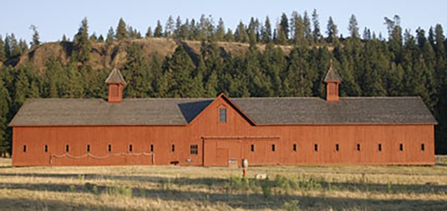 Long red barn with several windows. Black roof and two chimneys. Ponderosa pine covered hillside behind the building.