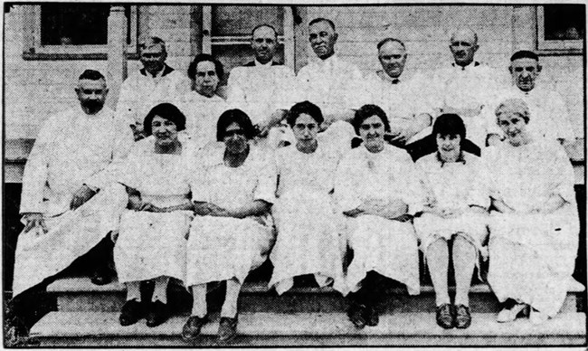 Grainy historic black and white newspaper photo of Doctors, physicians, and nurses sitting together on the outside steps of their rural hospital on an Indian Reservation