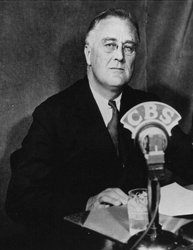 A black-and-white photo. A man sits behind several microphones labeled with the call letters of broadcast stations.