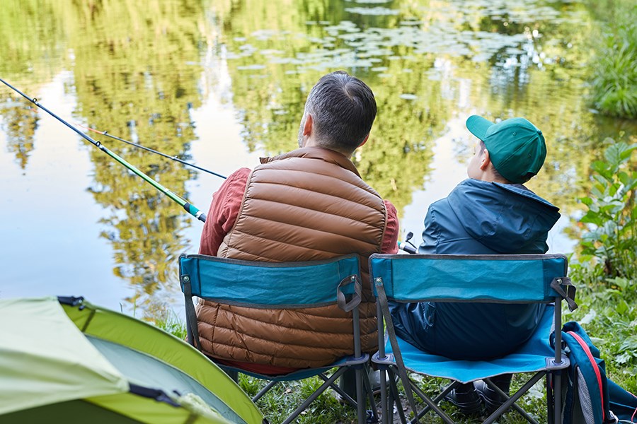 An adult and child sitting in camp chairs, fishing at the edge of a lake.