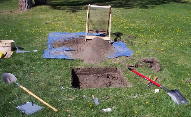 Archeology tools, screen, shovel, blue tarp around a square hole.