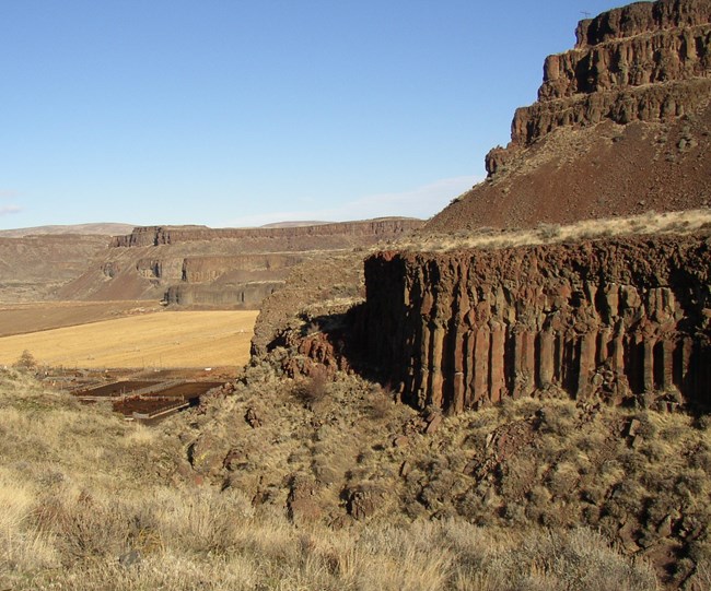 A landform with layers of dark rock in the foreground, more rock and farmland in the background