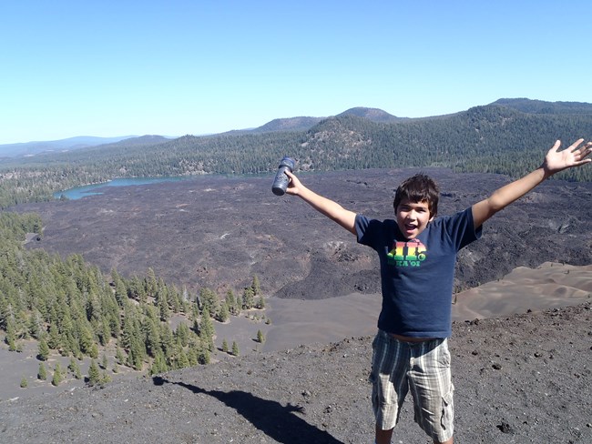 a boy faces the camera and lifts his arms over his head in excitement at the cinder cone behind him