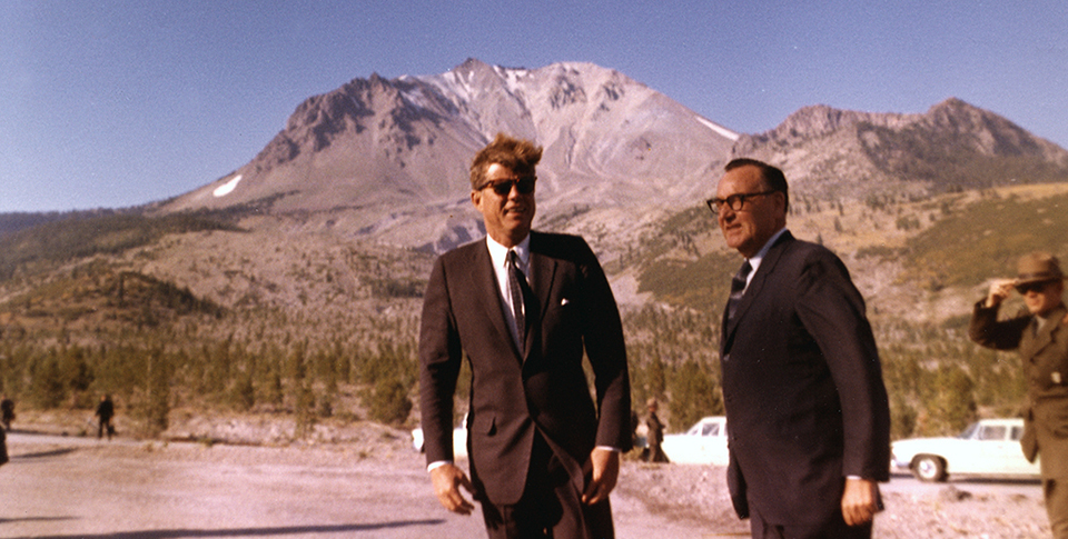 Two men in black suits standing, backed by a bare volcanic peak. A ranger in a green uniform holds his hat down in the background at right.