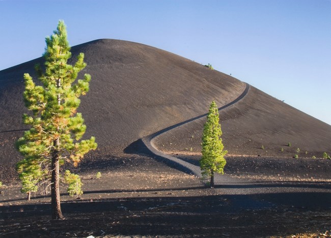 a large conical dome of loose gray material looms in the background, a winding trail leading up to the summit