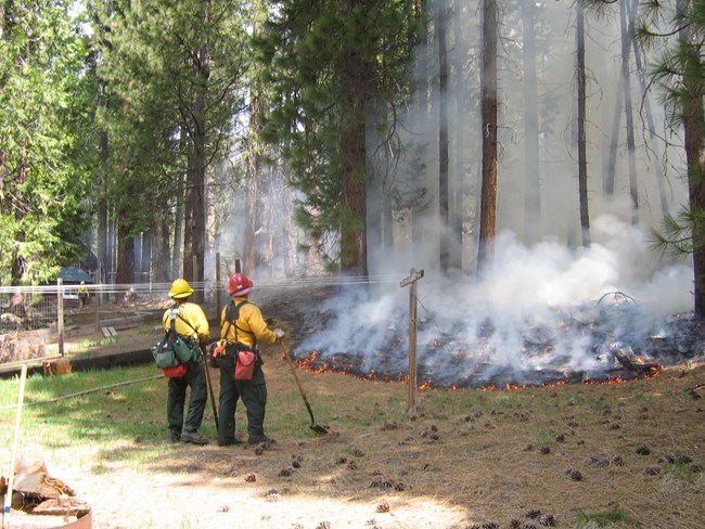 two firefighters lean on shovels as they watch over a burning woodpile