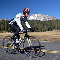 A bicyclist rides on the park highway with a meadow and snow-covered peak in the background
