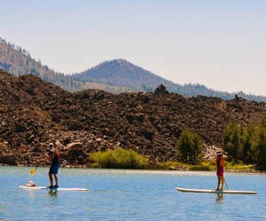 Paddle boarders on a blue lake edged by dark lava and backed by volcanic peaks.
