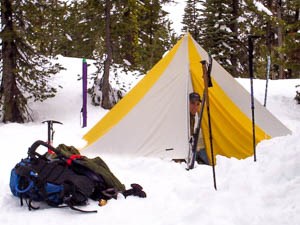 A man peeks out of a white and yellow tent on snow with two backpacks and ski gear nearby.