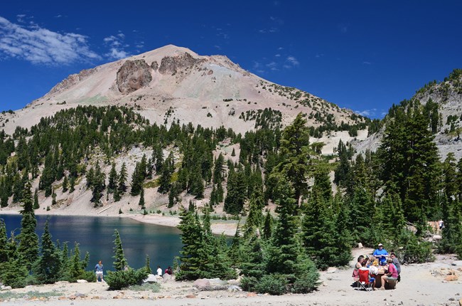 Picknickers sitting on a bench near Lake Helen with Lassen Peak in the background.