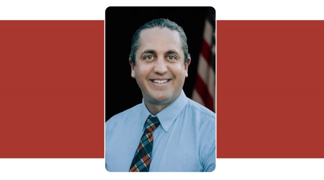 Portrait of man with hair pulled back and wearing shirt and tie. American Flag background.