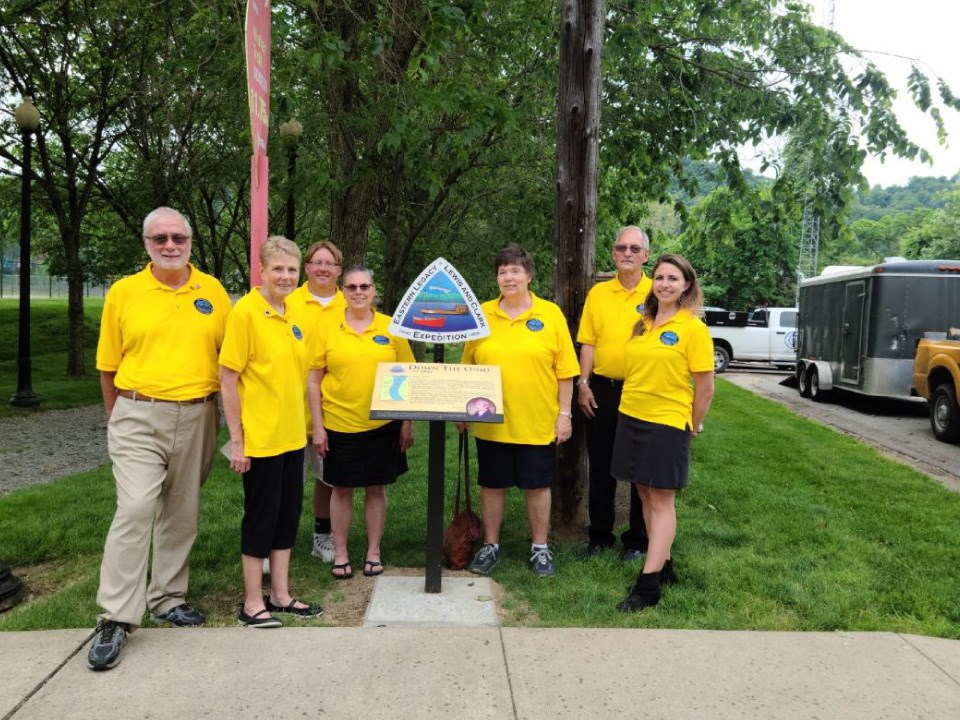 7 people in matching yellow shirts stand around sign. Insignia reads Eastern Legacy, drawing of two boats. Plaque below with information and photos.