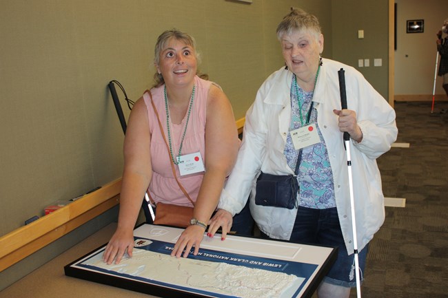 Two women smile as they feel a tactile map of the Lewis and Clark Trail. They hold white canes.
