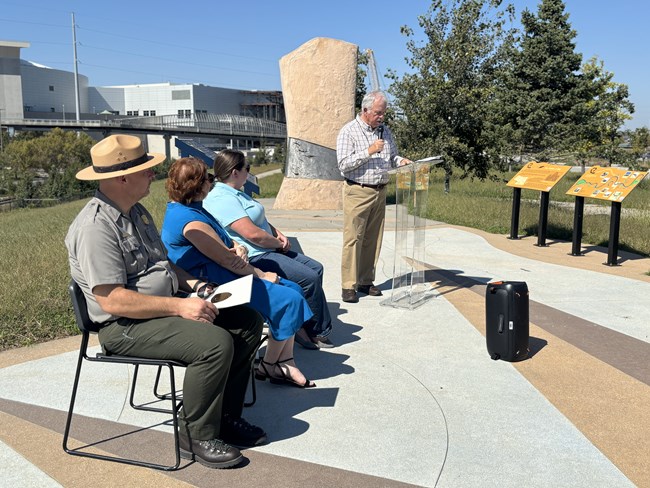 a group listens to a man speak at a podium outdoors