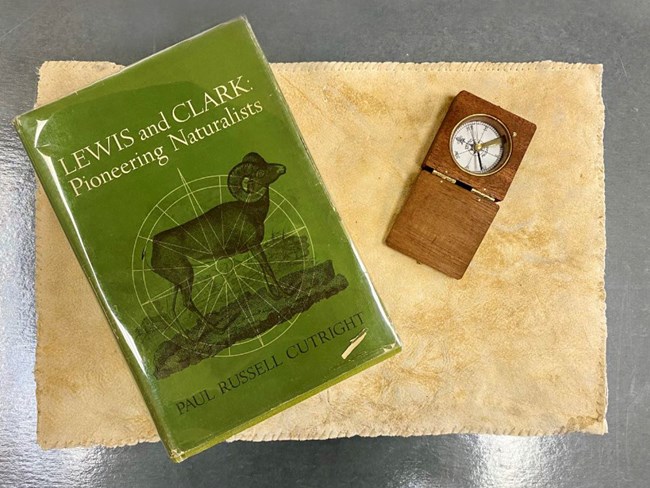 Small green book cover reads Lewis and Clark Pioneering Naturalists. Drawing of bighorn sheep. Leather placemat below.