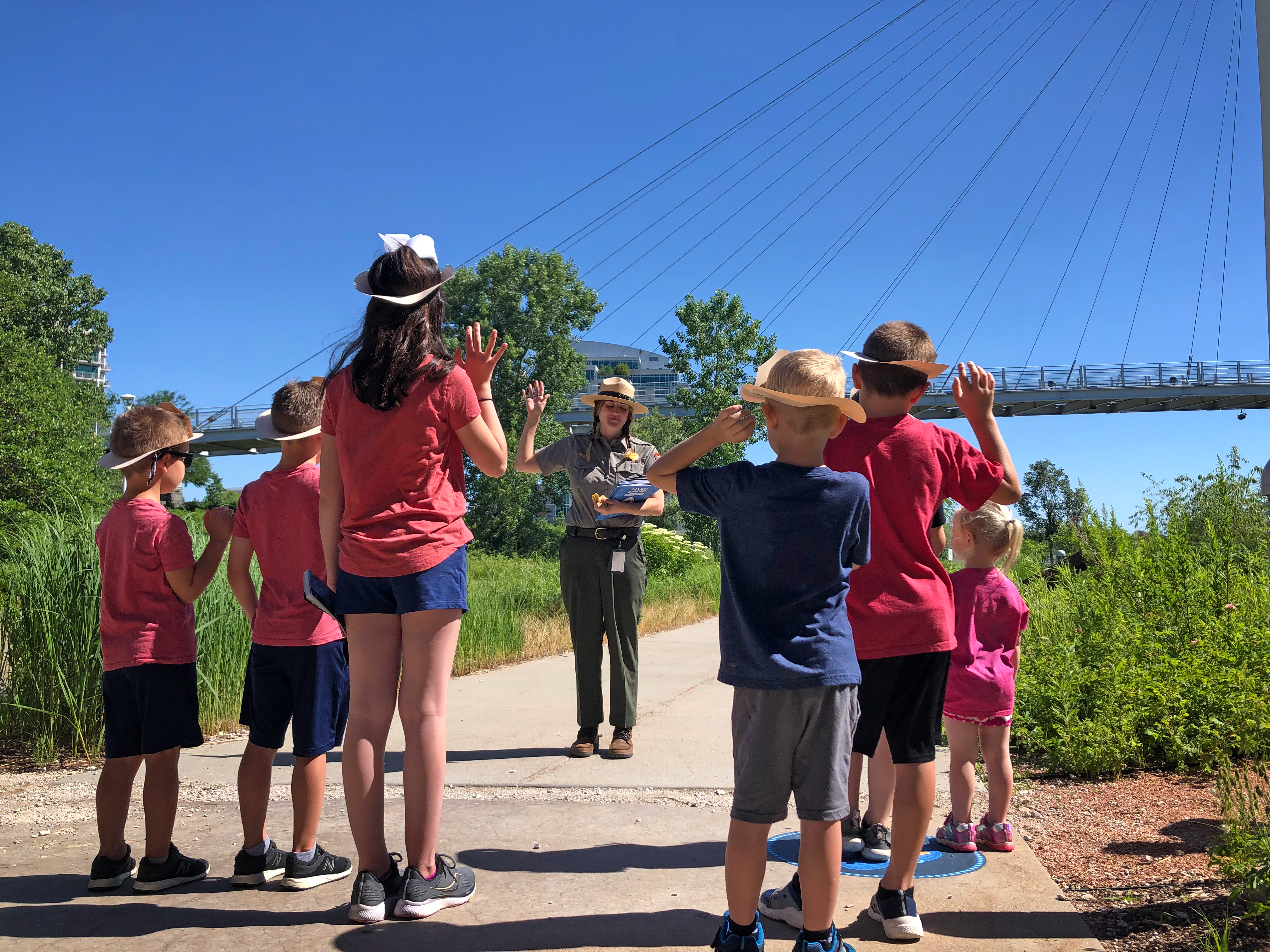 Ranger leads group of children in Junior Ranger Pledge. All stand wit their right hands raised. Bridge in background