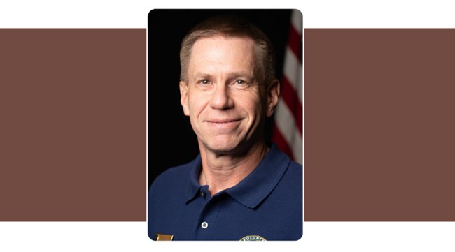 Portrait of man in blue polo with NPS Volunteer logo. American Flag background