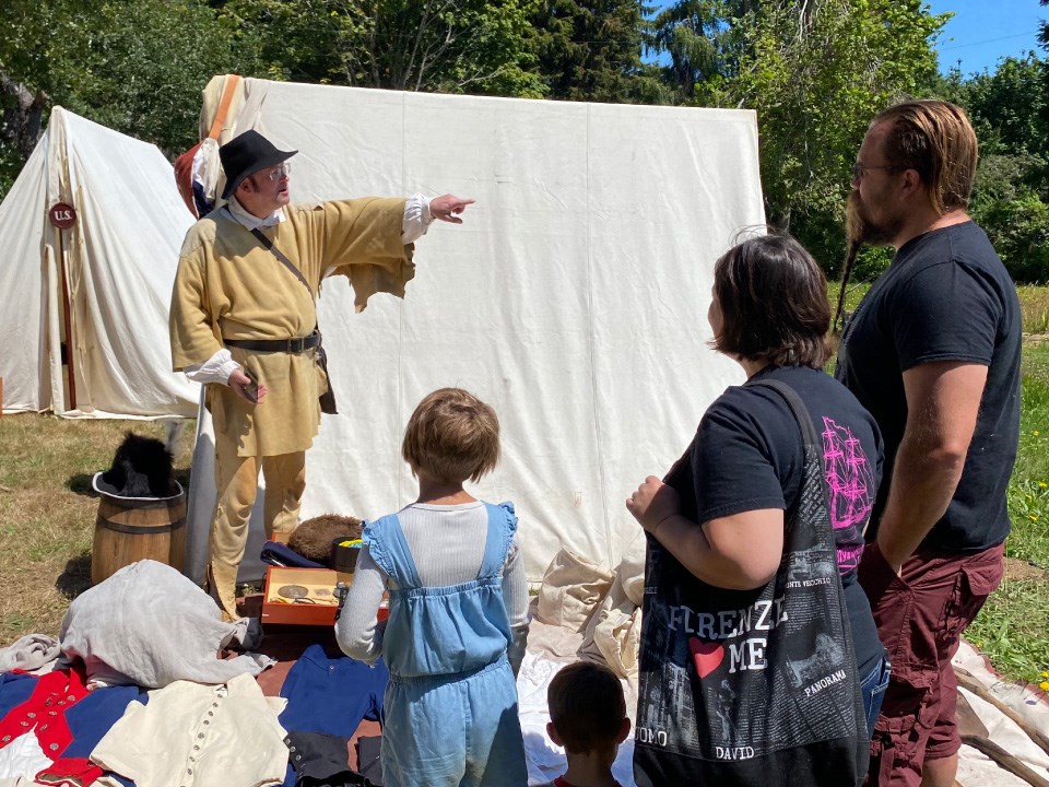 Man in early 1800s clothing address family of four. He points to the right. Canvas tent behind. Military uniforms and props laid out.