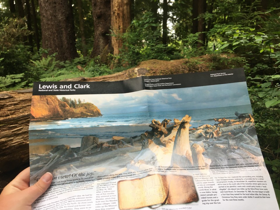 Hand holds up brochure with the National Park Service's iconic branding, black band on top. Brochure reads Lewis and Clark and features photo of rocky beach. Lush dark forest background.