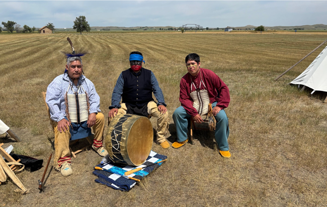 three men wearing Native American regalia sit outside with a large wooden drum