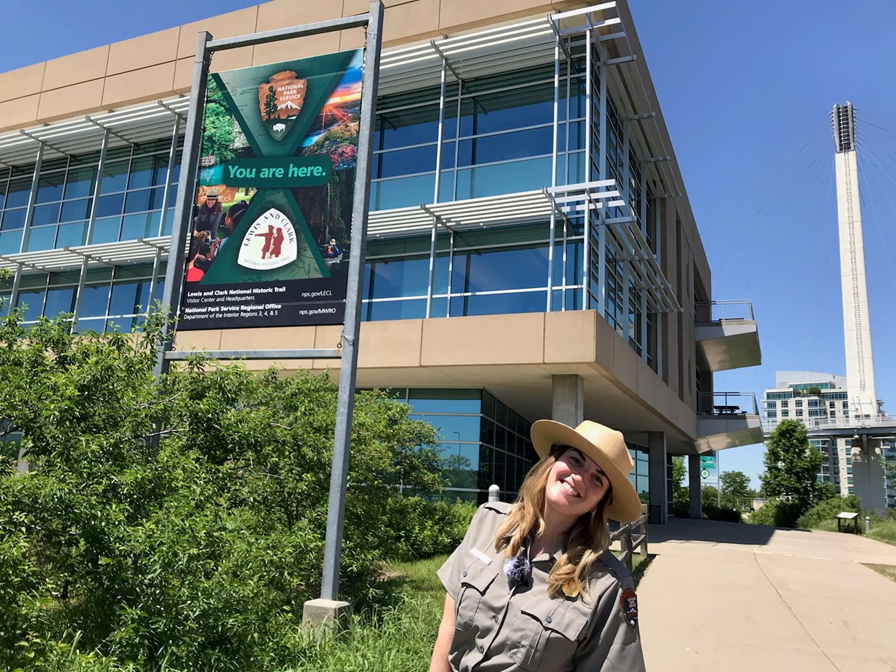 Ranger in uniform smiles in front of large office building. Banner behind has National Park Service and Lewis and Clark Trail logo