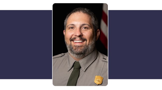 Man with short brown hair and short beard smiles broadly, wears Park Service uniform. American flag background,