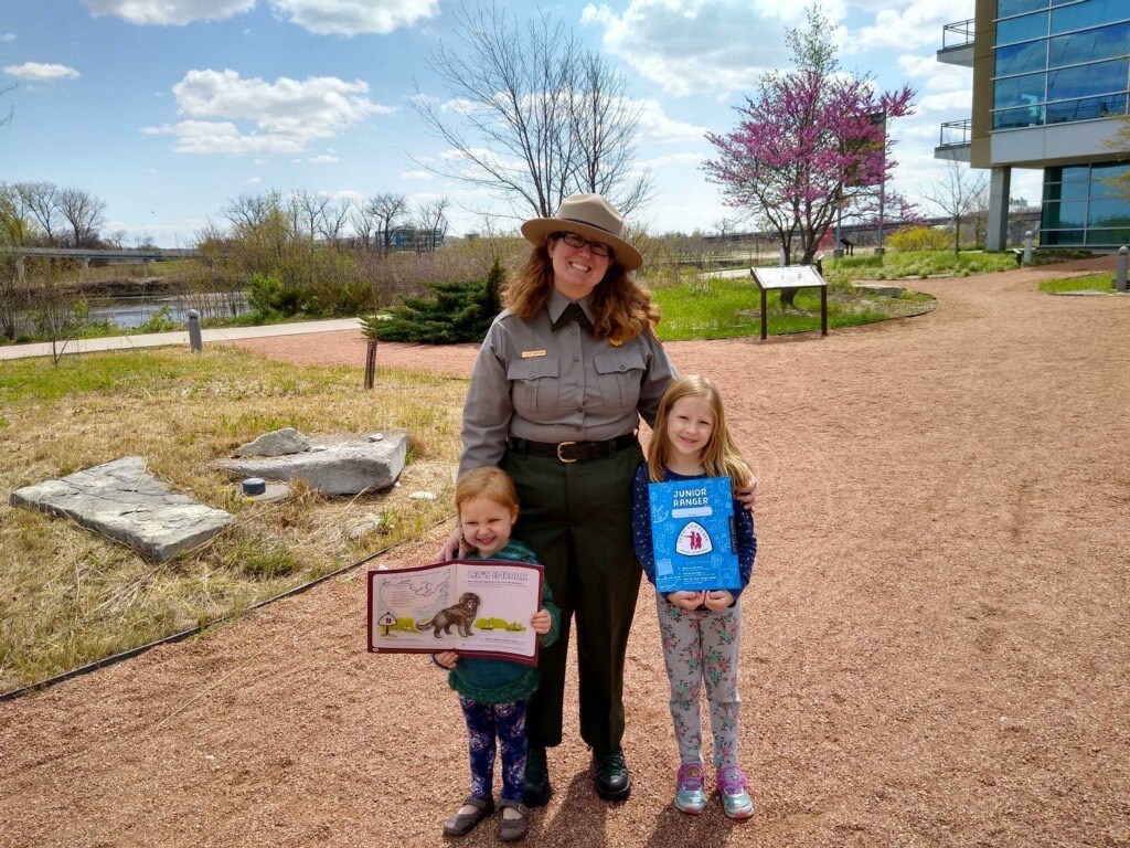 Ranger poses with two young kids who hold junior ranger booklets. River behind.