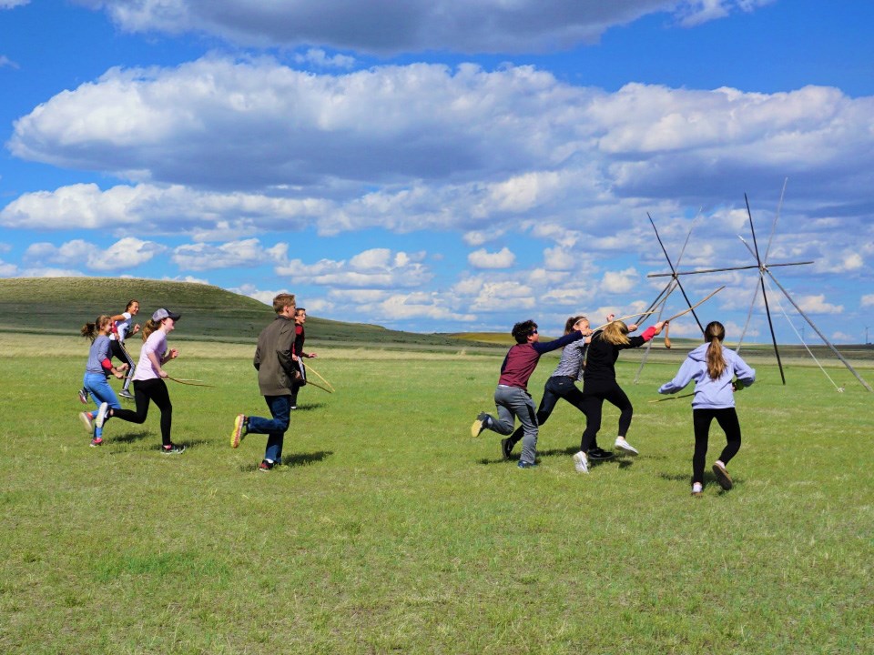 A group of teenagers run to the right holding sticks up in a game. Large grassy hill and blue sky. stick frame of teepee beyond.