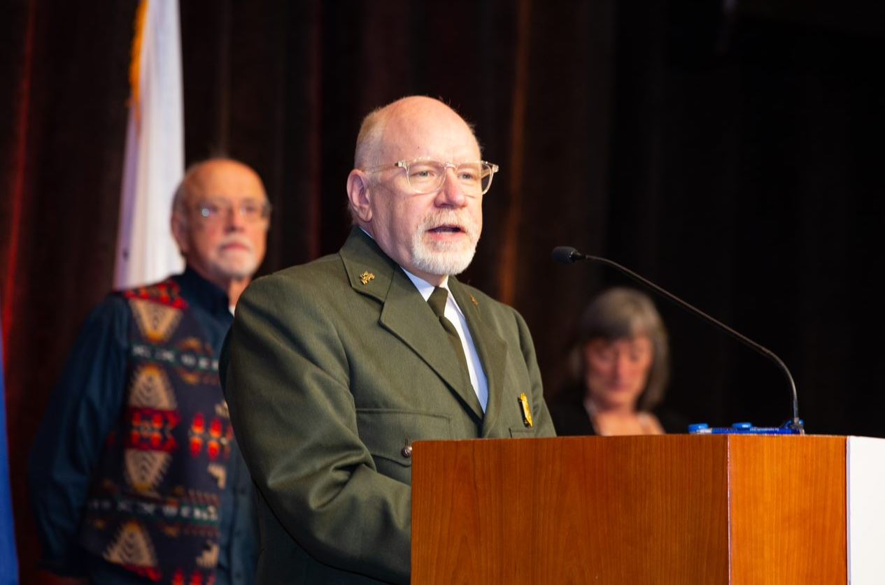 Man in National Park Service green formal jacket stands at podium and addresses audience.