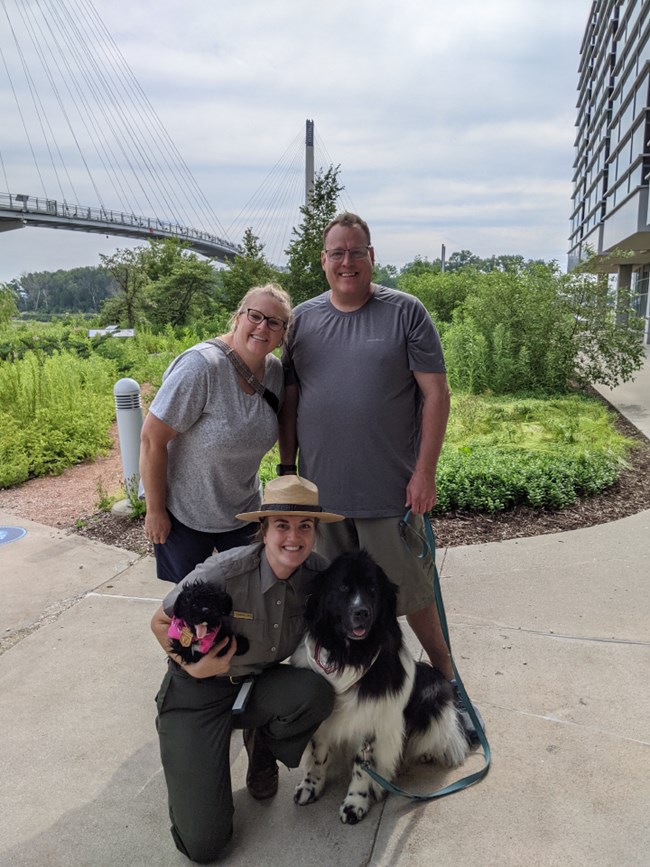 Two visitors and their dog pose with ranger at urban park. Dog is a black Newfoundland. Ranger holds black dog stuffed toy.