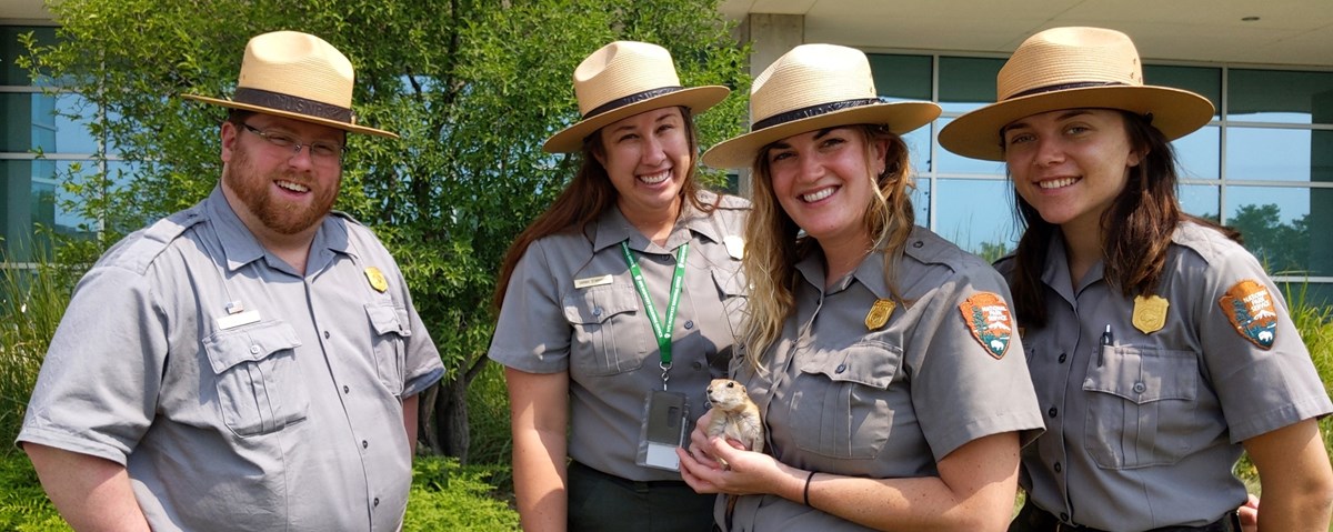 4 rangers in National Park Service Uniforms smile. One holds a black tailed prairie dog.