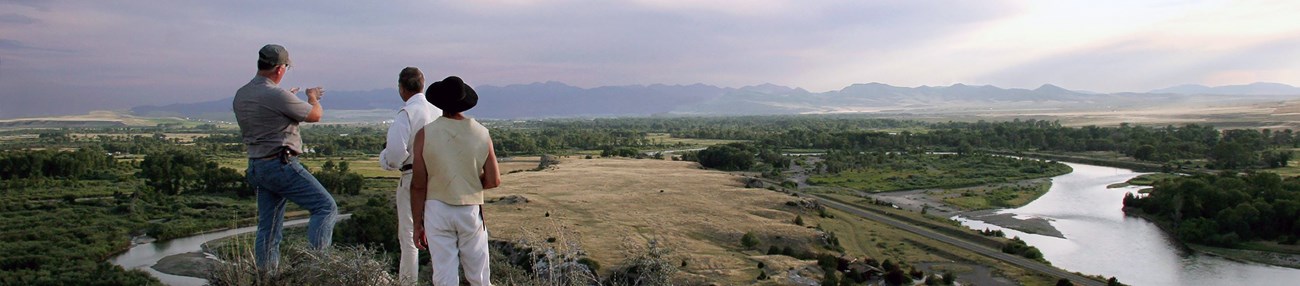 Three people overlooking a landscape with trees, mountains, and a river