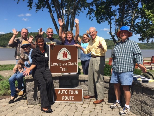 Eight people smile and wave, grouped around Lewis and Clark Trail Auto Tour Route Sign.