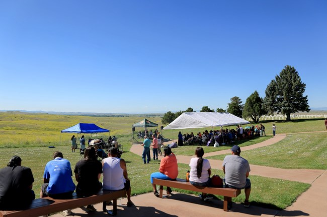 Groups of people sit under tents and on benches listening to speakers next to a cemetary with rows of white markers and a broad open field