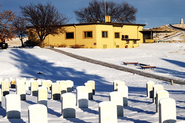 Winter landscape with white markers in the foreground, a tan building in mid-ground, and a monument in the background.  Snow covers the ground