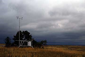 Weather station on the plains, in the fall,  sitting in front of a large green juniper tree.  The sky above is grey, blue, and white showing a storm moving in.